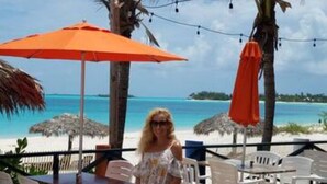 A beachside patio with white plastic chairs and tables, palm trees, and an orange umbrella.