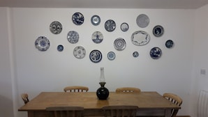 Un coin repas avec une table et des chaises en bois, un vase posé sur la table, et un mur décoré de différentes assiettes bleues et blanches.