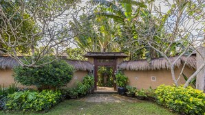 Um jardim com uma entrada de telhado de palha, plantas em vasos e um caminho que leva a uma construção.