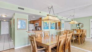 Un coin repas avec une table à plateau de verre et des chaises en bois, une cuisine à l’arrière-plan et un couloir menant à une porte.