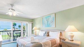 Une chambre d’hôtel avec un lit, deux lampes de chevet, une table de nuit, un balcon avec des chaises et un tableau au mur.