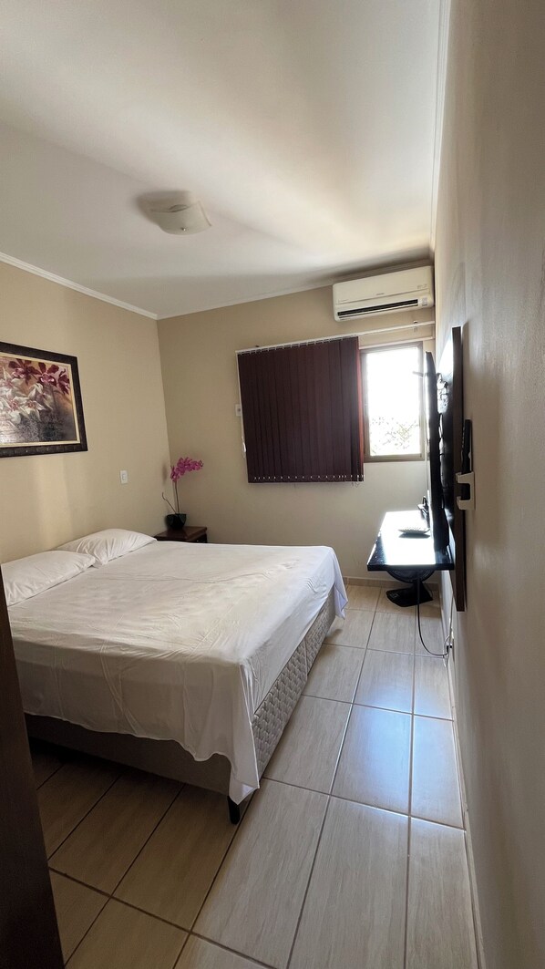A hotel room with a bed, a desk, a chair, a TV, and an air conditioning unit.