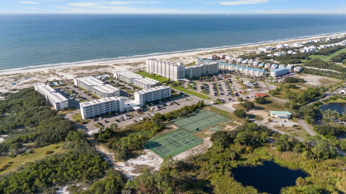Vista aérea de un complejo hotelero frente al mar con varias edificaciones, una cancha de tenis y una playa al fondo.
