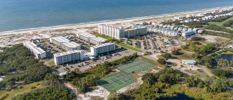 Vista aérea de un complejo hotelero frente al mar con varias edificaciones, una cancha de tenis y una playa al fondo.