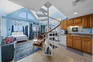 A spacious living area with a staircase leading to an upper level, a kitchen with wooden cabinets, and a large window offering a view. - Coastal Getaway | Sleeps 6 | Sport Courts, Community Pools, Beach Access (Gulf Shores)