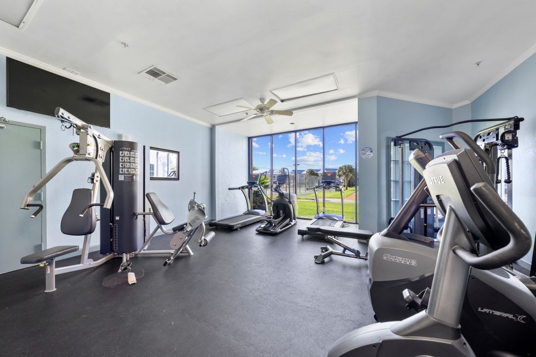 Ein Fitnessraum mit unterschiedlichen Trainingsgeräten, großen Fenstern und einem Deckenventilator.