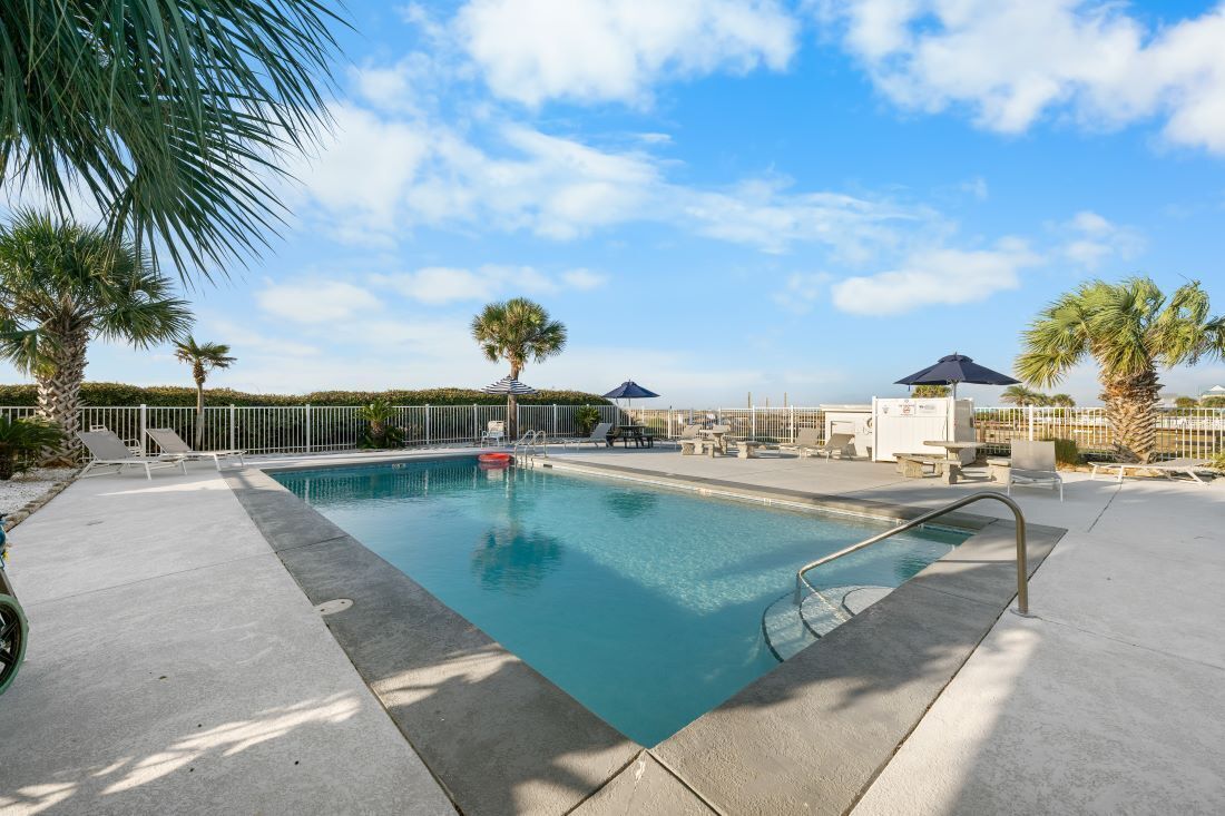 Ein Swimmingpool mit Liegestühlen und Sonnenschirmen, umgeben von Palmen und blauem Himmel.