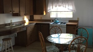 A compact kitchen with wooden cabinets, a gas stove, a sink, and a dining area with a table and chairs. - Rivers Edge/Off Grid/ Bird Watching Summer Paradise (Paradise)