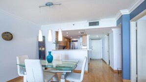 Un espace repas moderne avec une table en verre et des chaises blanches, un vase bleu, des luminaires suspendus et une cuisine en arrière-plan.