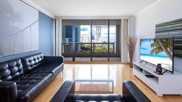 Un salon moderne avec un canapé en cuir noir, une table basse blanche et un téléviseur à écran plat fixé sur une console blanche. De grandes fenêtres offrent une vue sur le paysage urbain et une plage.
