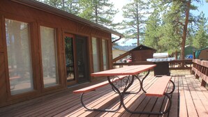 Eine Holzterrasse mit Picknicktisch und Grillplatz, umgeben von großen Fenstern und Blick auf Bäume.