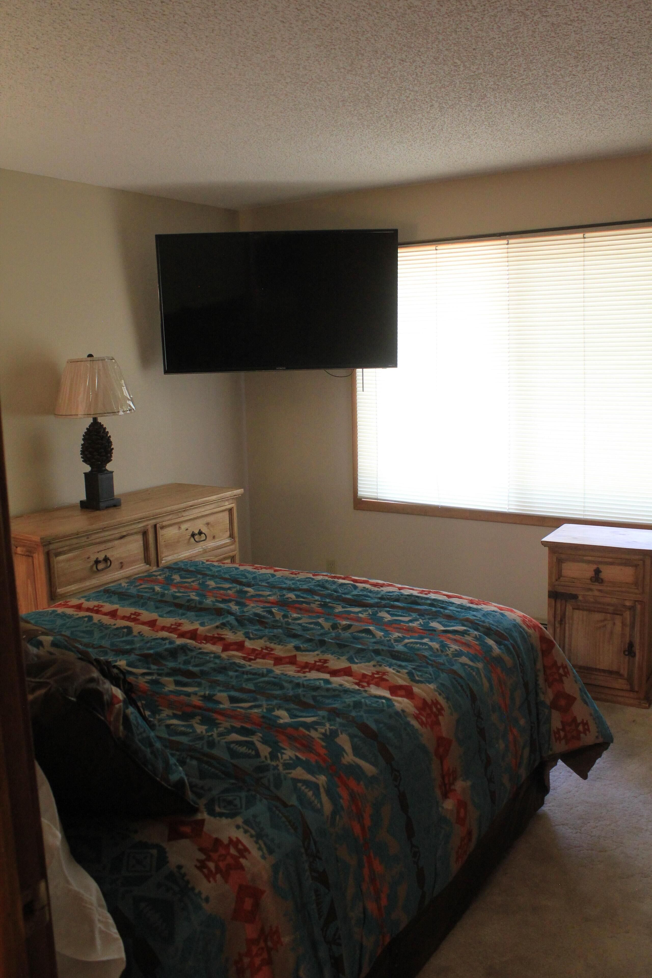 A bedroom with a bed, a dresser, a nightstand, a television mounted on the wall, and a window with blinds.