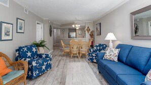 Een woonkamer met een blauwe bank, rieten stoelen, en een eethoek met een tafel en stoelen.