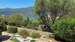 A landscaped garden with a stone pathway, various shrubs, and a view of a body of water and hills in the background.