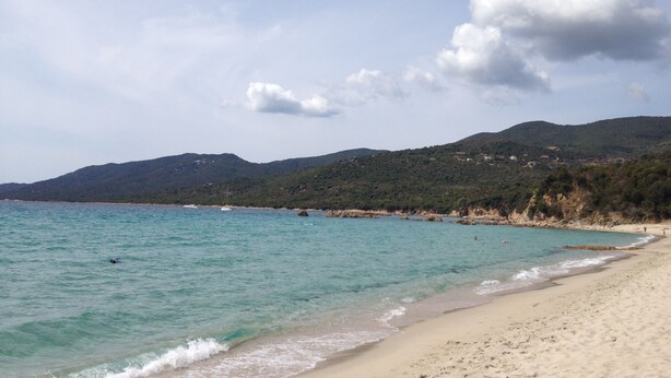 A beach with clear turquoise water, sandy shore, and a hilly background.