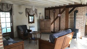 A cozy living room with a sofa, armchair, and a small table. There is a window with curtains, a radiator, and a brick wall with wooden beams. - Beautiful bressane house 4/5 people (Le Tartre)