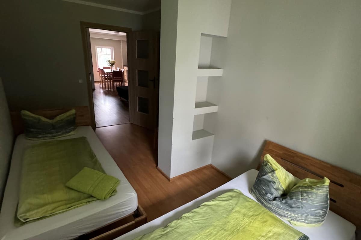 A room with two beds, each with green bedding and pillows, wooden headboards, and a white wall with a built-in shelf.