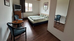 Une chambre d’hôtel avec un plafond en bois, un lit recouvert d’une courtepointe à motifs verts et blancs, une petite table avec une chaise, un téléviseur posé sur un meuble en bois et une fenêtre avec des rideaux.