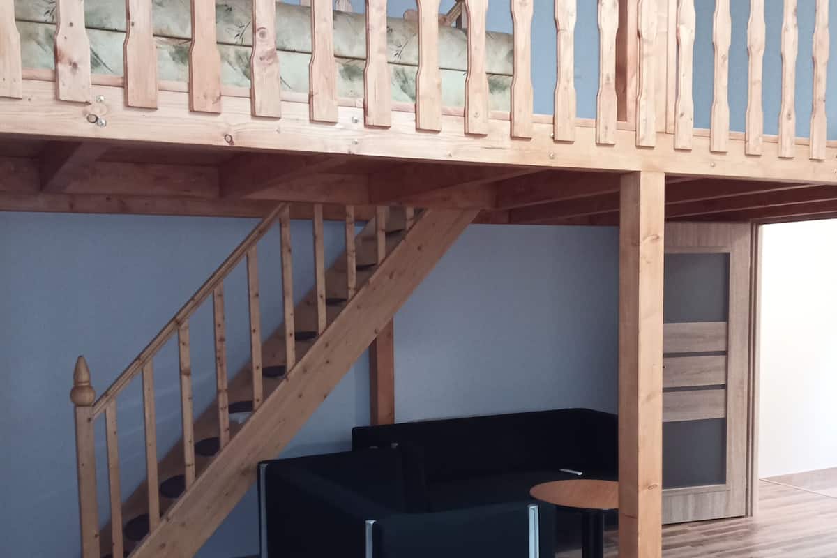 A room with a loft bed, a wooden staircase, a black sofa, and a small table.