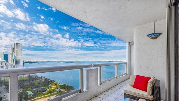 A balcony with a view of a city skyline and water. There is a sofa with a red cushion and a small table. - Bayfront Long-Stay | Premium Amenities | StayCozy (Miami)