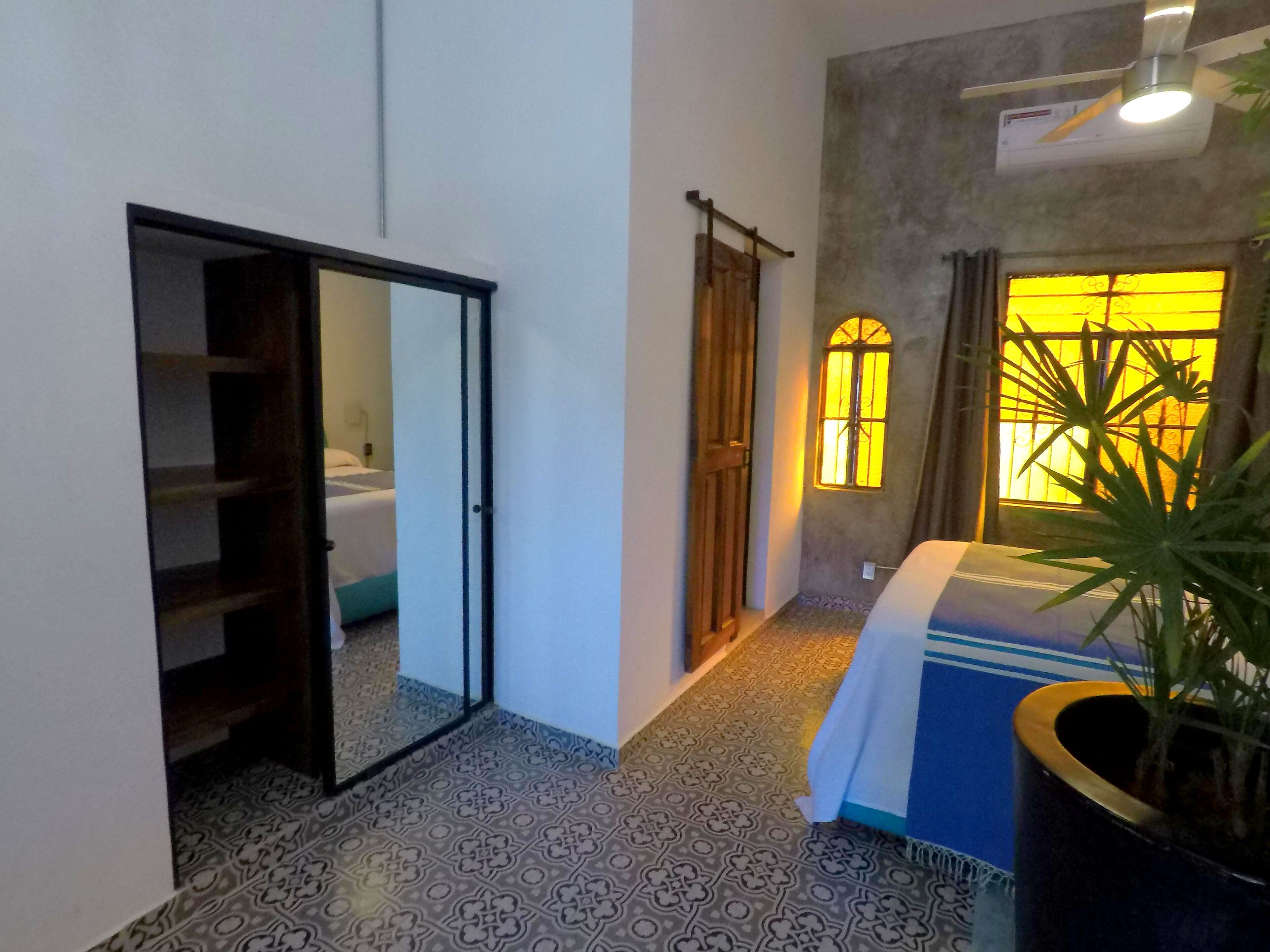 A room with patterned floor tiles, a bed with a blue and white striped bedspread, a potted plant, and a window with a yellow-tinted glass.