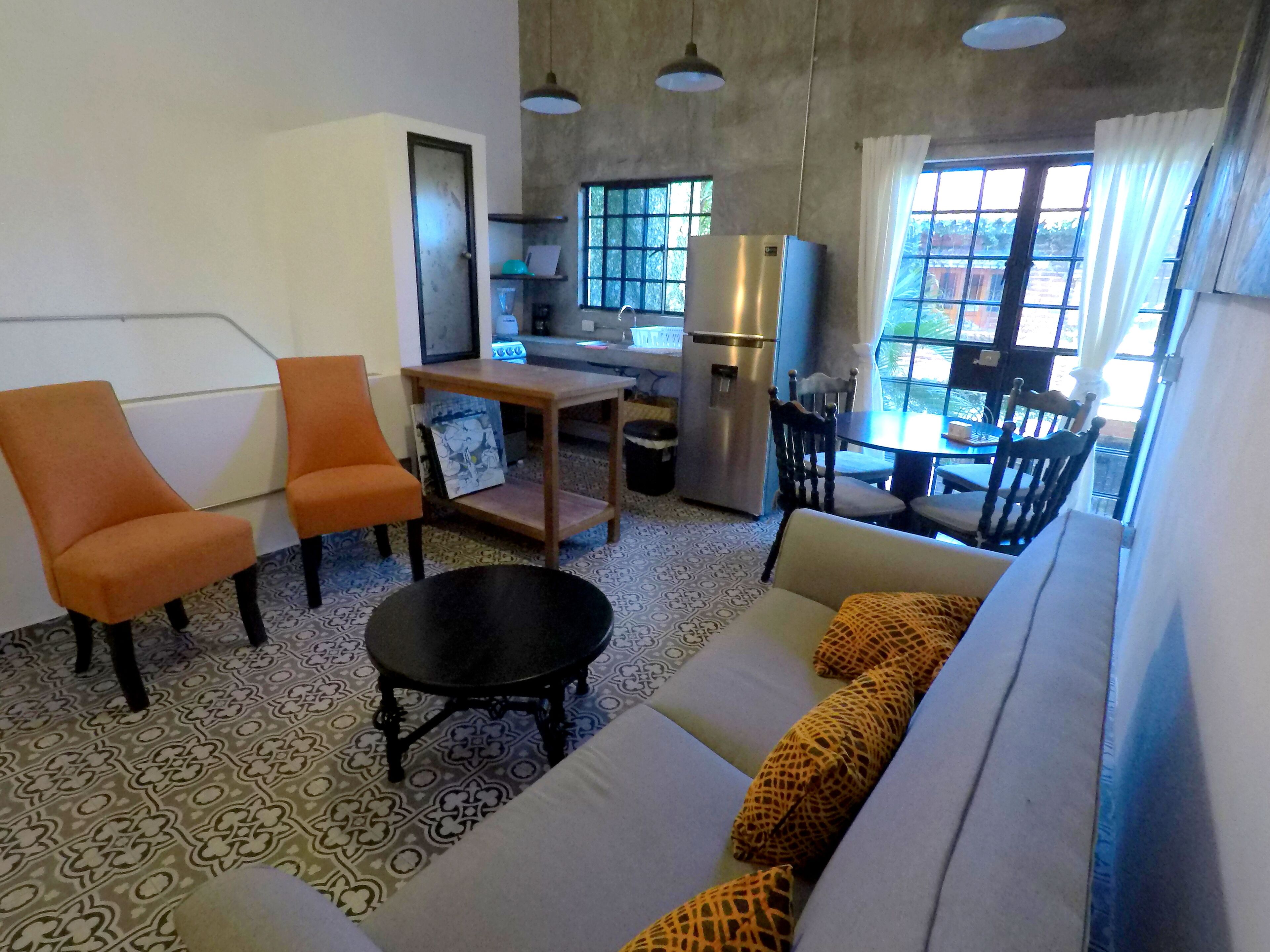 A compact living space with a sofa, two orange chairs, a small table, a black coffee table, a refrigerator, and a dining area with chairs and a table.