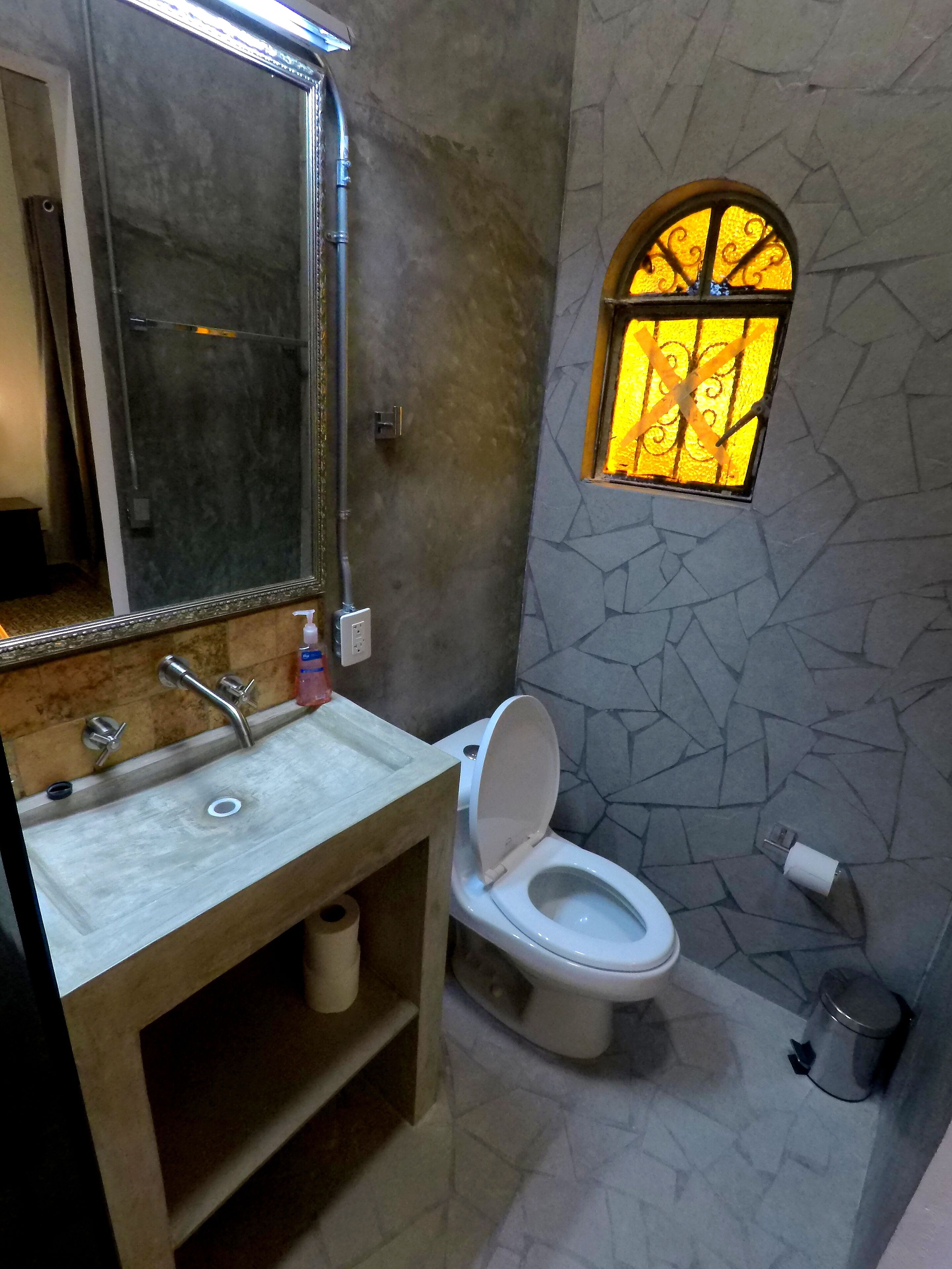 Un baño con lavamanos de mármol, un espejo, un inodoro, una ventana pequeña con un diseño de vitral y un cubo de basura.