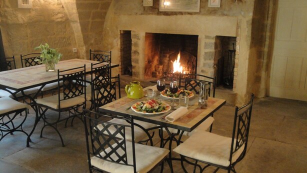 Zona pranzo con camino in pietra, tavolo apparecchiato per due con piatti di cibo e bicchieri di vino, e un vaso di fiori.