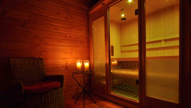 A wooden cabin interior with a sliding glass door leading to a sauna area. There is a wicker chair with a red cushion and a small table with candles. - Gîte "L'Anglin" in 160 ha estate with heated swimming pool, Spa-Sauna  (Chalais)