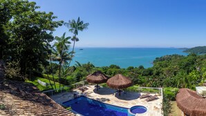 Zona de piscina con vista al océano, rodeada de exuberante vegetación y sombrillas de paja.