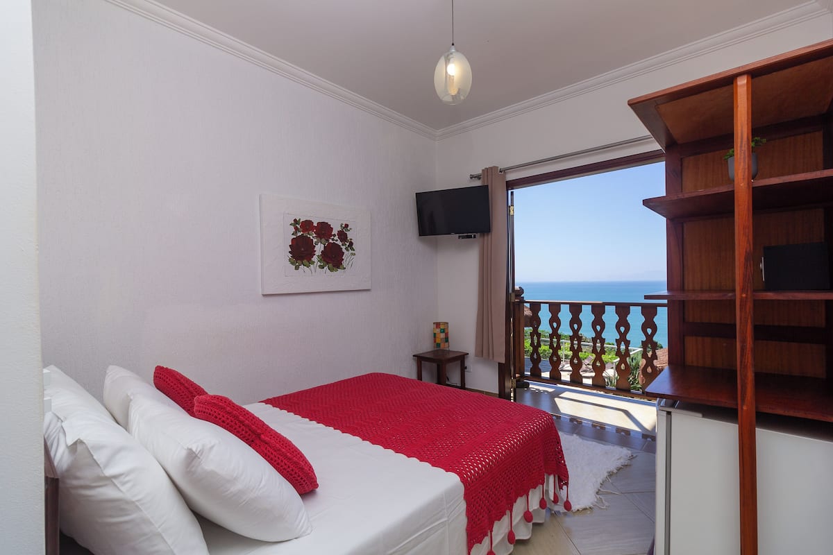 Une chambre à coucher avec un lit, une télévision, une couverture rouge et une vue sur la mer.