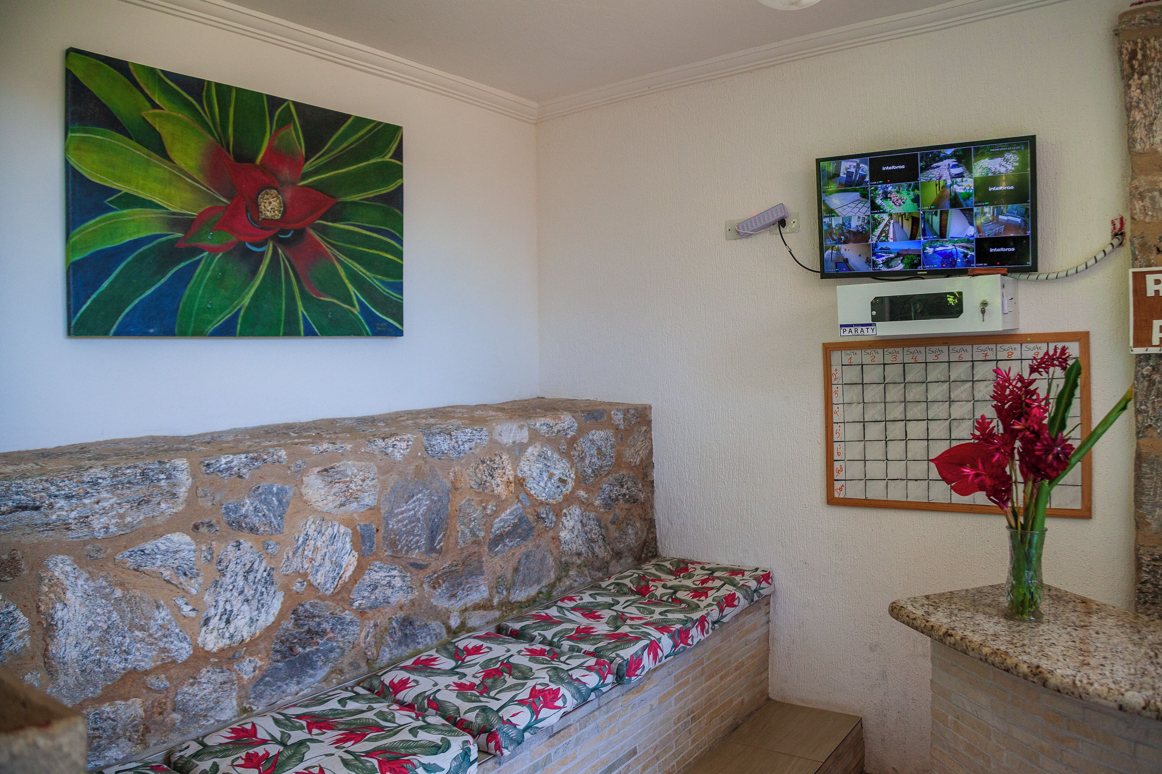 Eine Steinsitzbank mit floralem Bezug, ein an der Wand montierter Fernseher und eine Vase mit roten Blumen auf einer Anrichte.