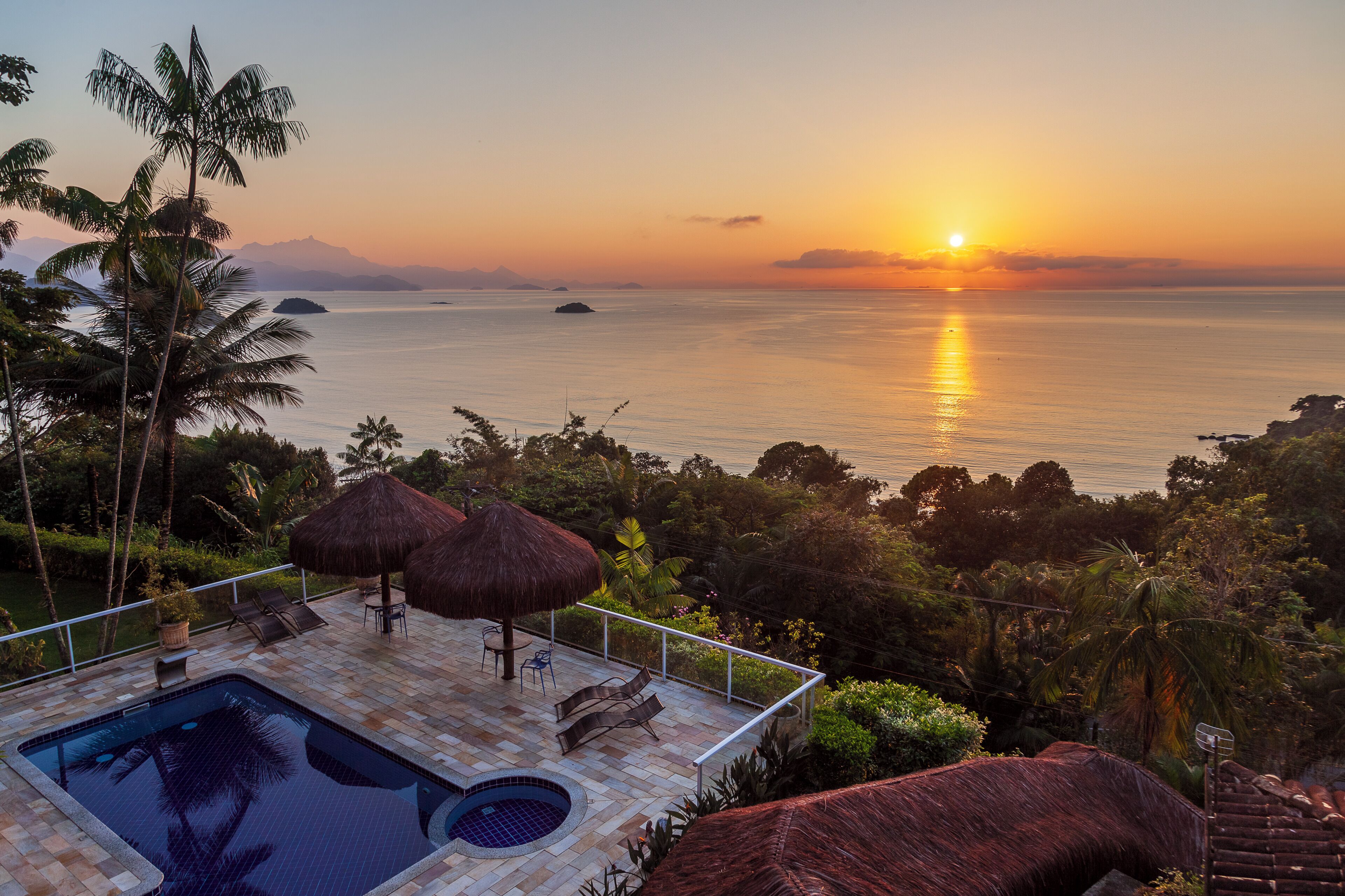 A pool area with a view of the ocean at sunset, surrounded by palm trees and a thatched roof structure.