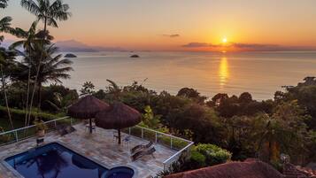 A pool area with a view of the ocean at sunset, surrounded by palm trees and a thatched roof structure.