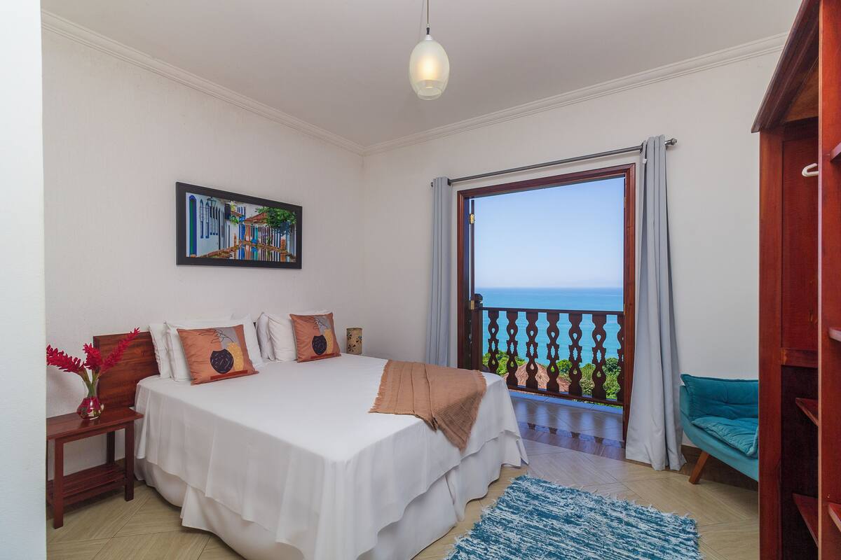 Une chambre à coucher avec un lit, une table de chevet, un vase de fleurs, un tableau et un balcon donnant sur la mer.