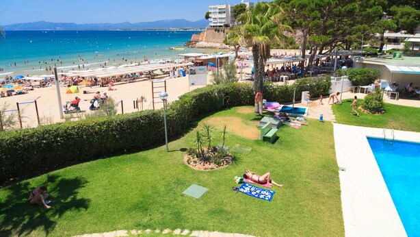 Ein Strandhotel mit Pool, einer Rasenfläche mit einer Palme und Blick auf das Meer und die Berge.