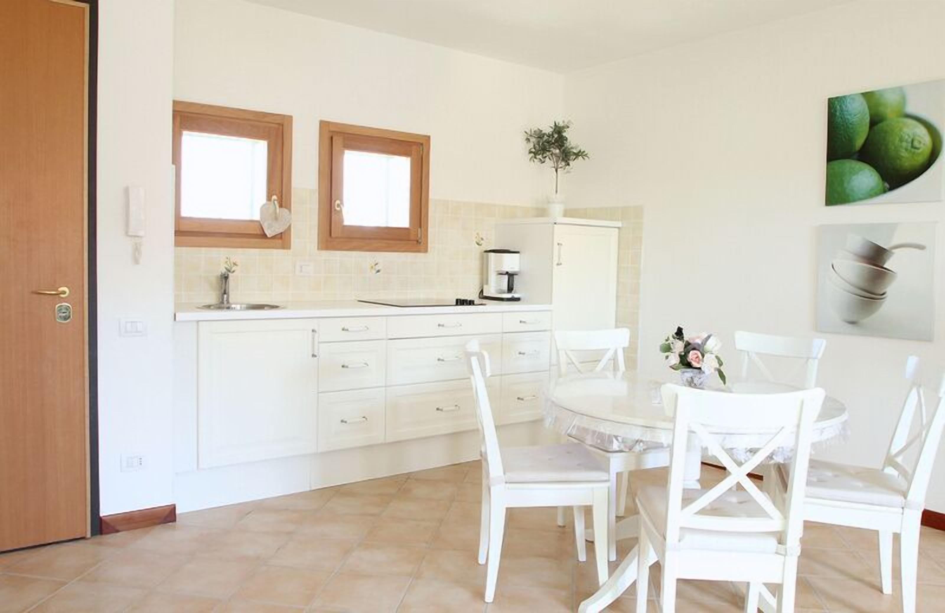 A modern kitchen with white cabinetry, two windows, a dining table with white chairs, and a small potted plant.