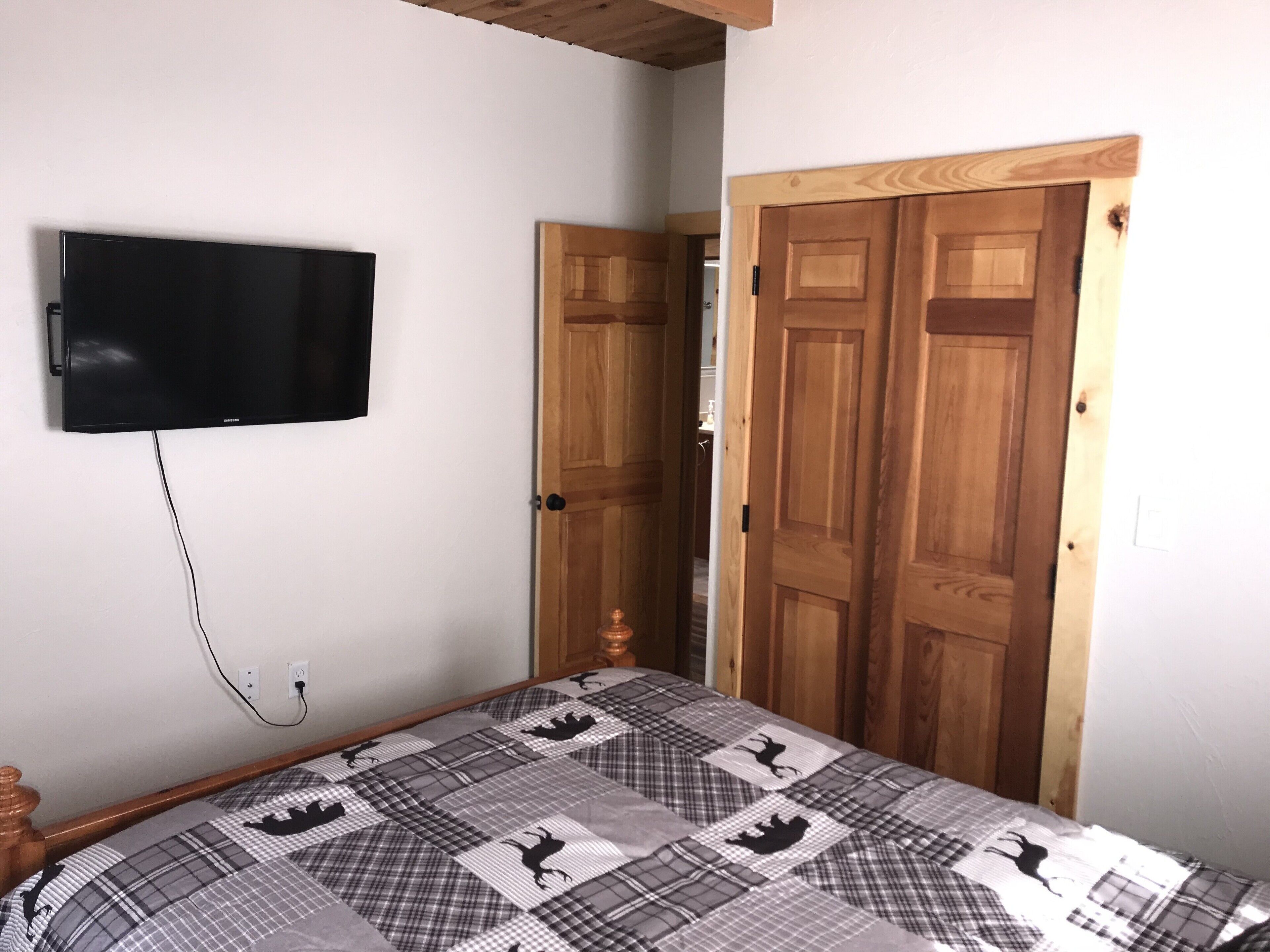 Um quarto com cama, televisão na parede e porta de madeira.