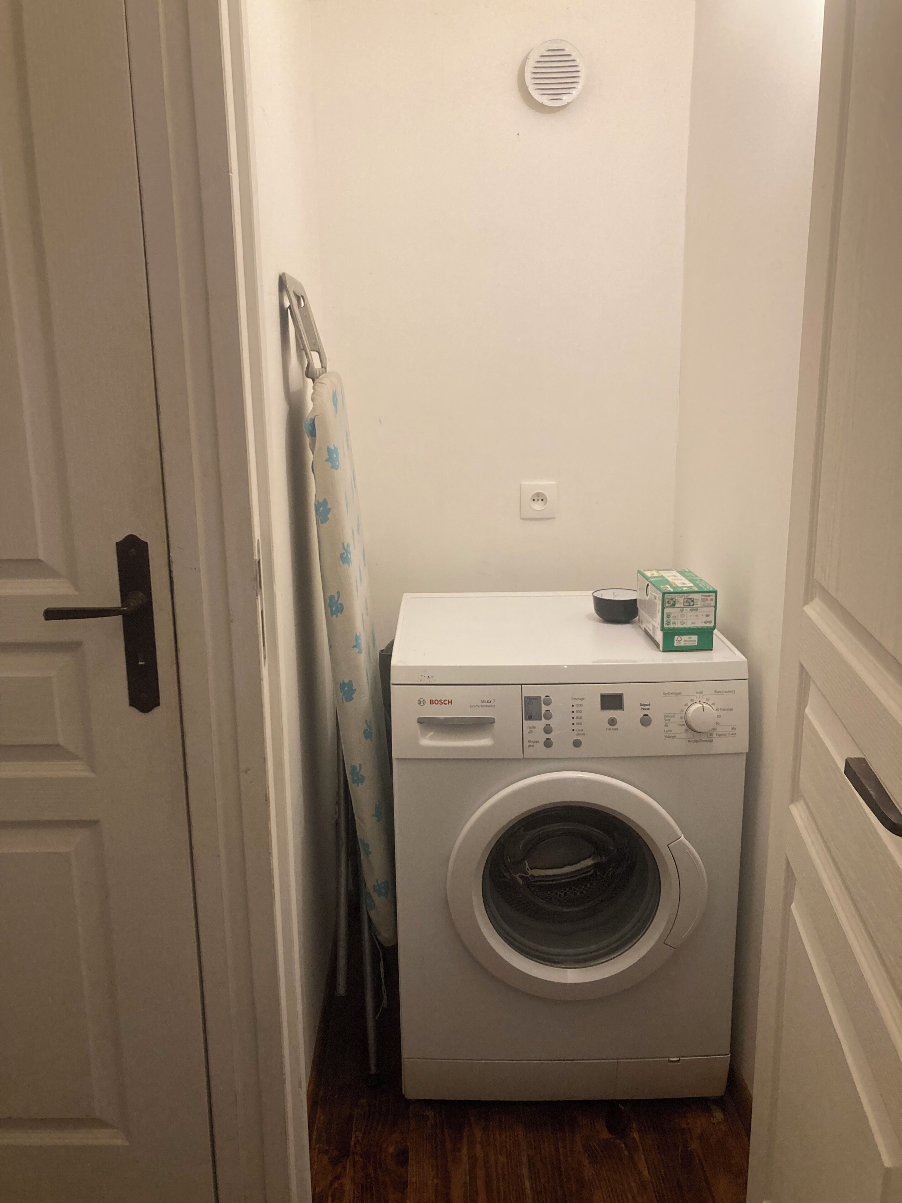 A compact laundry room with a Bosch washing machine, a drying rack, and a small box on top.