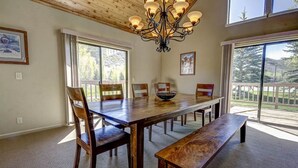 A dining area with a large wooden table, benches, and chairs. There is a chandelier above the table, and the room has large windows with a view of the outdoors.