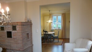 A living room with a brick fireplace, a dining area with a table and chairs, and a white armchair. - Lovingly furnished new FEWO for 2 people in an absolutely quiet location (Horn-Bad Meinberg / Feldrom)