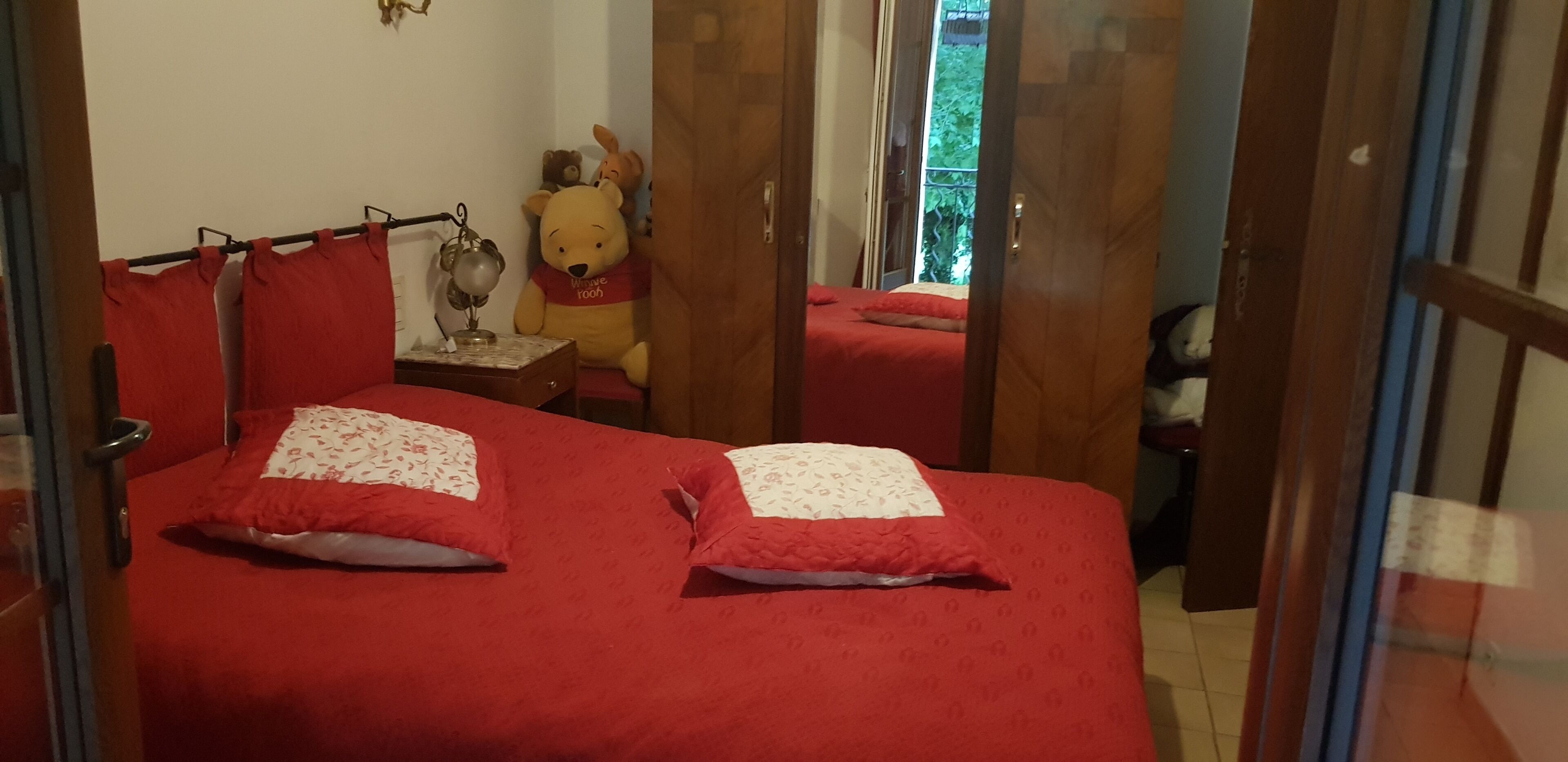A bedroom with two beds, each with red bedding and floral-patterned pillows. There is a nightstand with a lamp and a teddy bear. A window with curtains is visible in the background.