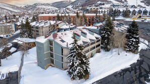 Un immeuble à plusieurs étages, dont le toit est recouvert de neige, entouré d’arbres également enneigés et avec une chaîne de montagnes en arrière-plan.