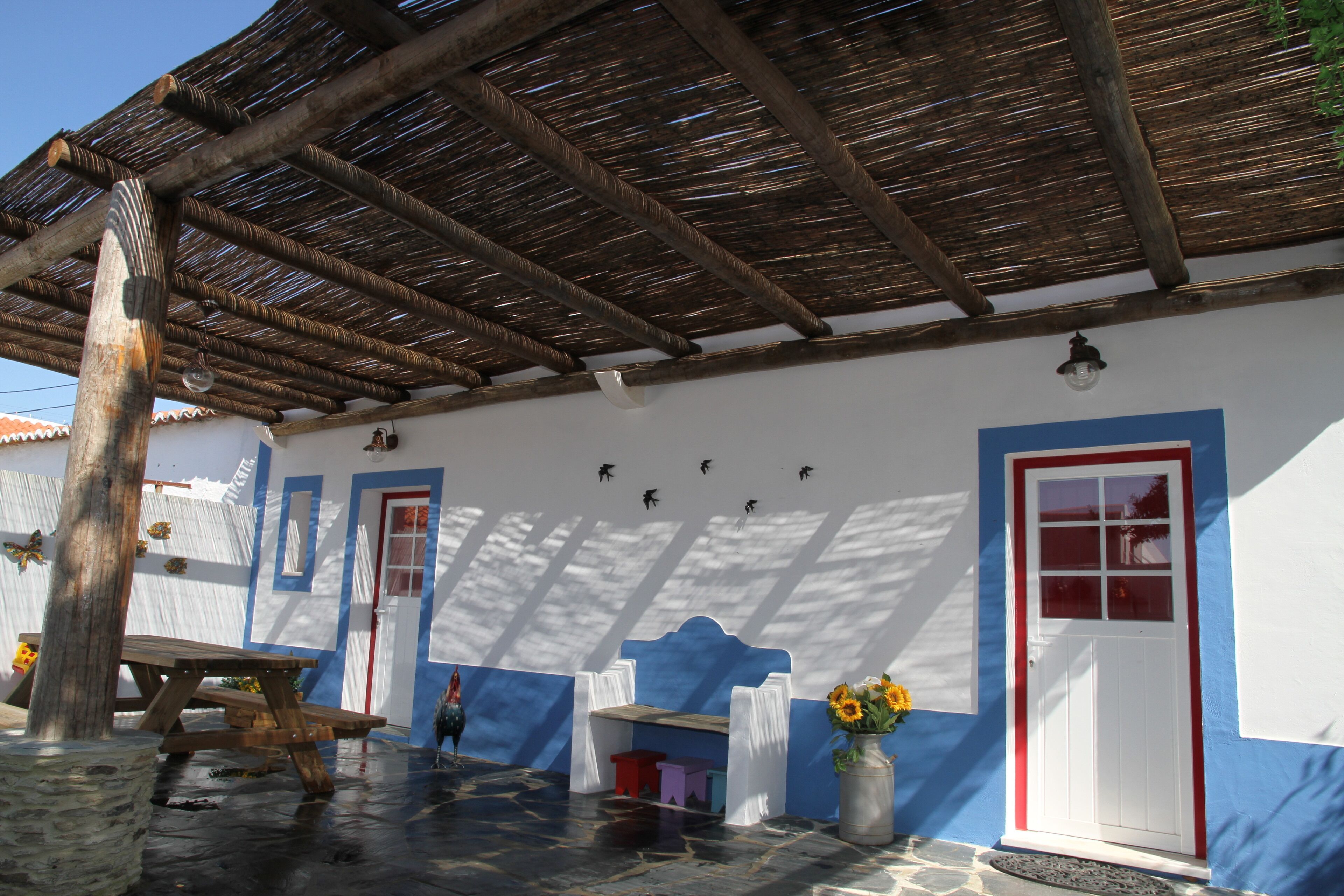 Un espacio exterior rústico con un banco de madera, una mesa, un taburete pequeño y un jarrón con flores.