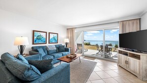 A modern living room with a sofa, coffee table, and a flat-screen TV. There are two framed pictures on the wall, a sliding glass door leading to an outdoor area, and a wooden cabinet with a TV stand. - Beach House 103D  - Ground Floor - Beachfront! (Miramar Beach)