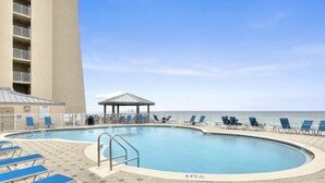 A swimming pool with lounge chairs and a gazebo, surrounded by a tall building and a view of the ocean. - Beach House 103D  - Ground Floor - Beachfront! (Miramar Beach)