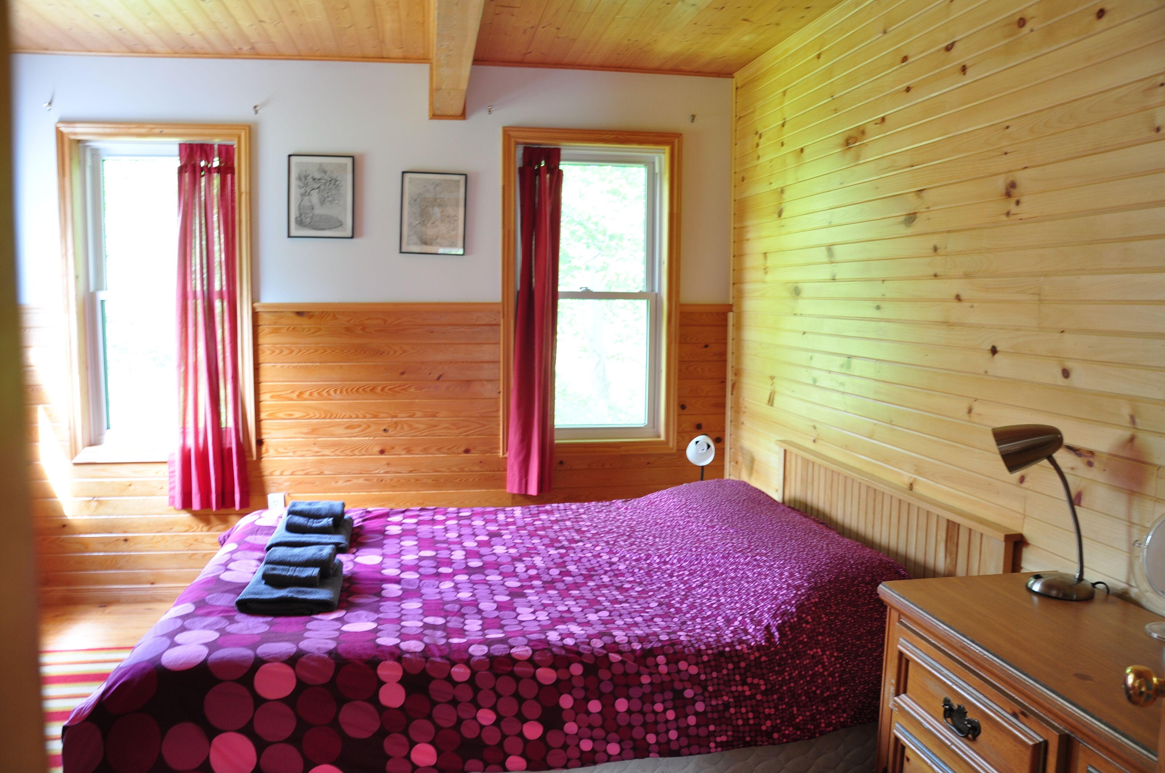 A room with wooden walls, a bed with a pink polka dot bedspread, two windows with curtains, a desk with a lamp, and a framed picture on the wall.