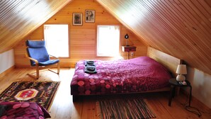 Un dormitorio acogedor en el ático, con techo inclinado de madera, una cama con colcha estampada, una silla azul y una mesita de noche con lámpara.