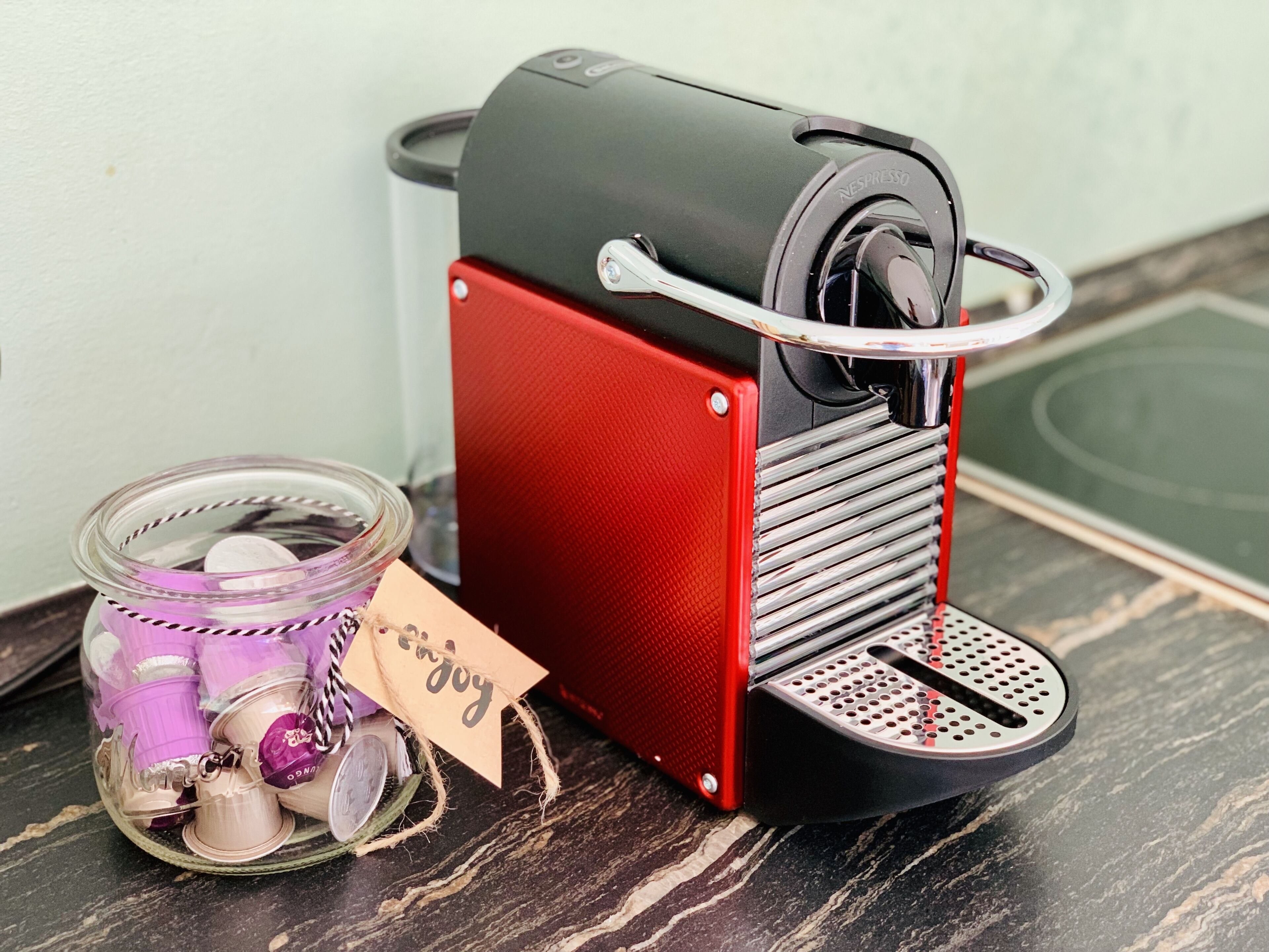 A coffee machine with a red and black design, a glass jar with tea bags, and a tag that reads "Enjoy".
