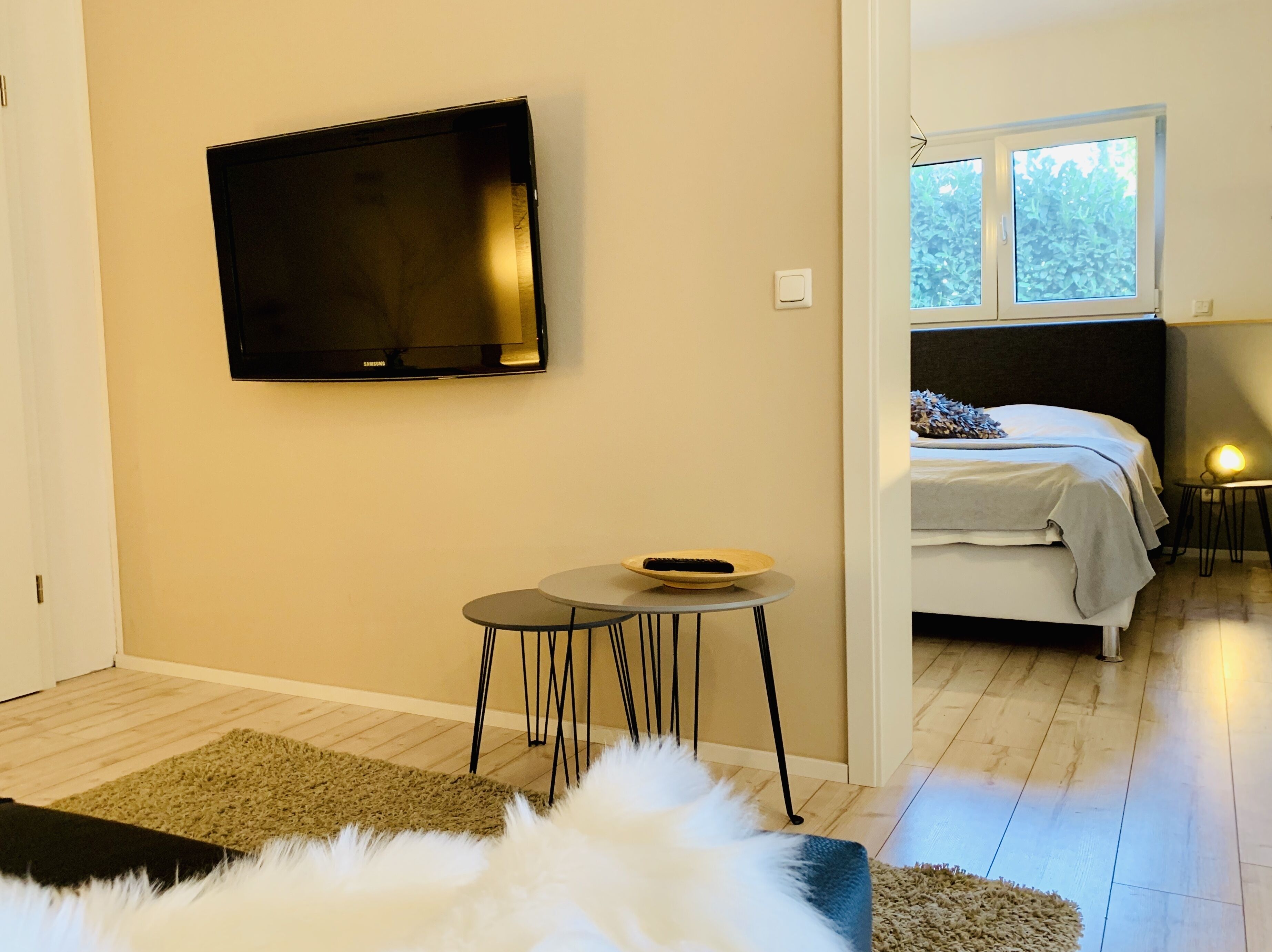 Une chambre moderne avec un lit, une petite table sur laquelle se trouve un bol, un téléviseur fixé au mur et une fenêtre donnant sur un paysage verdoyant.